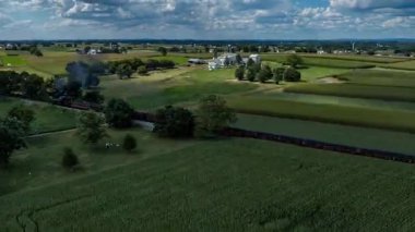 A hyper-Lapse of a steam train winds through lush green fields under a blue sky filled with clouds. The train is surrounded by crops and trees, showcasing the beauty of rural landscapes.
