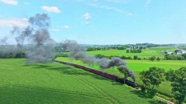 Paradise, Pennsylvania, 8-8-2025 - A historic steam locomotive chugs along a picturesque landscape filled with lush green fields and a clear blue sky. Smoke billows as it makes its way through the