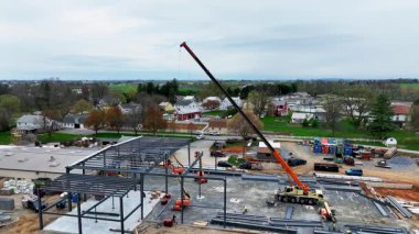 Bir şantiye, faaliyetlerini koordine eden işçilerle doludur. Yeni bir yapı inşa etmek için büyük bir vinç malzemeleri kaldırıyor. Gökyüzü bulutlu bir günde evler ve tarım arazileriyle çevrilidir..