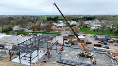 Büyük inşaat alanı çelik kirişleri kaldıran bir vinç gösteriyor. Yapılar kırsal kesimde şekillendikçe işçiler aşağıda koordinasyon içinde çalışırlar. Bölgeyi çevreleyen yeşil alanlar.