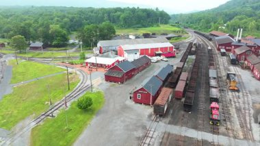 Bir hava manzarası, yeşil bir manzaraya yerleştirilmiş tarihi bir tren istasyonunu sergiliyor. Raylar, renkli binalar ve park edilmiş tren vagonları ile tüm alana yayılmış durumda..