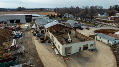 İşçiler kırsal bölgedeki bir inşaat alanındaki bir yapıyı yıkıyorlar. Kamyonlar ve ekipmanlar yakında ve gri gökyüzünün altında yeni evler görülebiliyor..