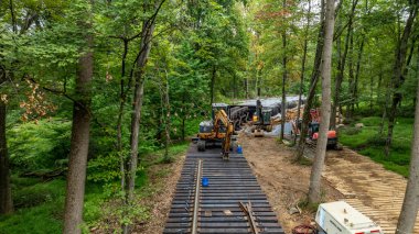 Yoğun makineler verimli bir ormandaki demiryolu inşaat projesinde çalışıyor. İşçiler açık gökyüzü altında ekipman taşıyorlar ve malzemeleri yönetiyorlar. Etrafını saran ağaçlar, doğayı gözler önüne seriyor..
