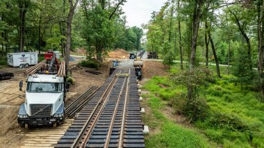 Ağaçlarla çevrili bir demiryolu hattında inşaat çalışmaları sürüyor. Rayların yakınına bir kamyon ve makineler yerleştiriliyor ve ek malzemeler de yakınlarda görünüyor..