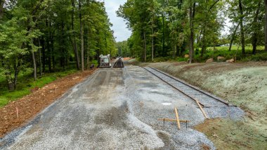 İşçiler orman ortamında yeni demiryolu rayları boyunca çakıl döşüyorlar. Her iki taraftaki ağaçlar, sonbaharın başlarında olduğunu gösteriyor. Makineler görünür, devam eden inşaatı gösteriyor.