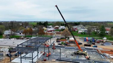 İşçiler küçük bir kasabadaki bir inşaat alanında meşguller. Çelik kirişler kuruyorlar ve yeni bir bina için zemin hazırlıyorlar. Yakın evler ve tarlalar bulutlu bir gökyüzünün altında görülebilir..