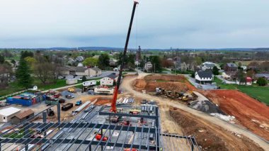 Kırsal kesimde bir inşaat alanı işçilerin malzeme kaldırmak için vinç kullandıklarını gösteriyor. Yakınlarda, arka planda manzaralı tepeler bulunan arazinin etrafında araçlar ve ekipmanlar bulunuyor..
