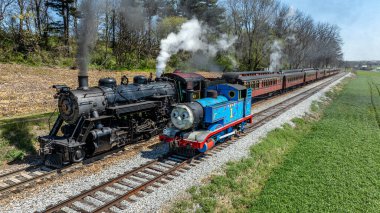 Cennet, Pennsylvania, 17 Nisan 2025 - Açık bir günde yeşil tarlalar ve ağaçlarla çevrili iki buhar treni paralel gidiyor. Bir tren klasik siyahtır, diğeri ise parlak mavi..