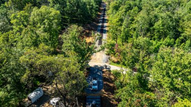 Hava perspektifi, ağaçlarla kaplı ormanın içinden geçen dolambaçlı bir patikayı ortaya çıkarır. Yol boyunca, sıcak güneş ışığıyla yıkanmış çeşitli yapılar görülebilir..