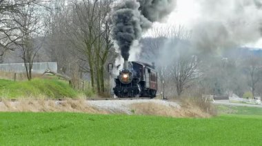 Antika bir buhar treni yeşil tarlalar ve çıplak ağaçlarla çevrili kırsal bir hat boyunca tıkırdıyor. Motorun sesi, havayı dumanla doldururken yankılanır..