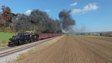 Ronks, Pennsylvania, 8 Kasım 2025 - Güzel sonbahar manzarasının ortasında klasik bir buhar treni raylar boyunca tıkırdıyor. Motorun dumanı ağaçların ve tarlaların yanından geçerken yükseliyor..