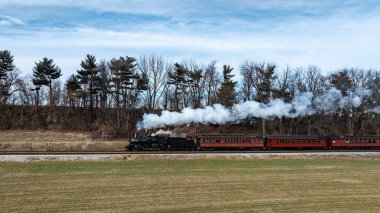 Eski bir buhar treni raylar boyunca tıkırdıyor, açık mavi gökyüzüne beyaz duman salıyor. Çevreleyen yeşillik ve ağaçlar kırsalda pitoresk bir bahar manzarası yaratıyor..