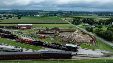 Ağır makineler, eski bir tren istasyonunda yeni bir döner tablonun döşenmesi üzerinde çalışıyor. Eski tren vagonları bulutlu bir gökyüzünün altında, kırsal manzaranın cazibesini sergiliyorlar..