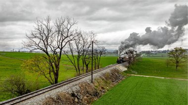 Gri gökyüzünün altında yemyeşil çayırlarla çevrili demiryolu rayları boyunca ilerleyen eski bir buhar treni. Motordan dumanlar yükseliyor. Resmedilmeye değer kırsal alanlarda seyahat ediyor..
