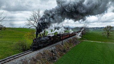 Cennet, Pennsylvania, 12 Nisan 2025 - Demiryolu rayları boyunca eski bir buhar treni, yemyeşil tarlaların ve bulutlu gökyüzünün zeminine yoğun dumanlar salıyor..