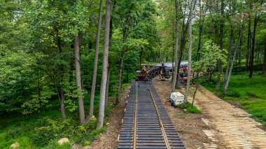 Ağır makineler ve inşaat ekipmanları bir orman açıklığında çalışmakla, ahşap demiryolu bağları kurmakla meşguller. Çevreleyen ağaçlar, mevsimi gösteren canlı sonbahar renkleri sergiliyor..