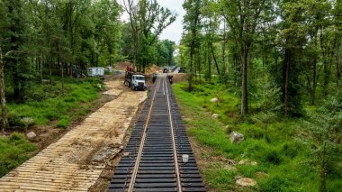 İşçilerin ormanlık bir alanda yeni demiryolu rayları döşediği bir inşaat alanı görülebilir. Bölgede hareket eden ekipmanlar ve rayları çevreleyen yeşil ağaçların görüntüsü yer alıyor..