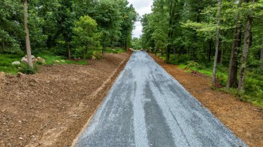 Yeşillik bir ormanın içinden geçen yeni bir çakıl yolu inşaatı devam ediyor. Ağaçlar yol boyunca uzanır. Erişilebilirliği ve güvenliği arttırmak için her iki tarafta da toprak temizlendi..