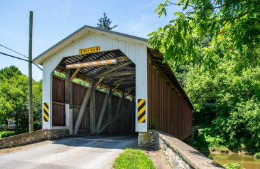 Ahşap kaplı bir köprü kırsal bir bölgedeki küçük bir nehri kapsar. Yapı ağaçlar ve yeşil manzarayla çevrilidir. Gün ışığının parladığı açık bir gün..