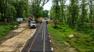 İşçiler yeşil, ağaçlık bir alanda yeni demiryolu rayları döşemekle meşguller. Ağır makineler mevcut ve inşaat sahasında parlak bir gökyüzü altında dağınık malzeme ve ekipmanlar yer alıyor..