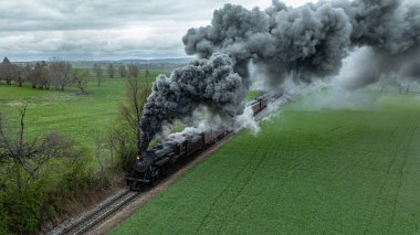 Bir buhar treni yeşil alanlar boyunca ilerliyor. Trenden dumanlar yükselirken hareket ediyor. Gökyüzü bulutlu, kırsal kesime yeni bir manzara ekleniyor. İnsanlar bu manzaranın tadını çıkarabilir..