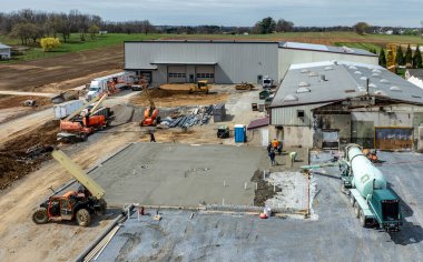 İşçiler, makineler ve araçlar kırsal alandaki bir inşaat alanında aktifler. Çalışmalar devam ederken ekip yeni bir binanın temelini atmak için beton döküyor..