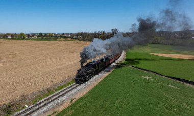 Bir buhar treni, yeşil tarlalar ve mavi gökyüzünün altındaki kahverengi tarım arazileriyle çevrili demiryolu rayları boyunca hareket eder. Motor ilerlerken duman dalgalanır..