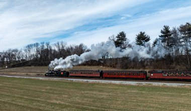 Bir buhar treni, motorundan yükselen dumanla raylar boyunca ilerler. Tren mavi gökyüzünün altında yeşil tarlaları ve çıplak ağaçları geçiyor. Sahne kırsal kesimde tipik bir günü gösteriyor..