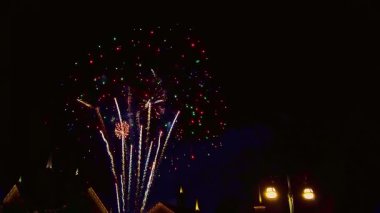 Şehir parkı yakınlarındaki bir gece kutlamasında binaların üzerinde parlak havai fişekler gökyüzünü aydınlatıyor. Kalabalık, renkli patlamaların havayı heyecanla doldurmasını izlemek için toplandı..