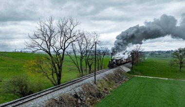 Klasik bir buhar treni, bulutlu bir gökyüzünün altında yemyeşil tarlalar ve çıplak ağaçlarla çevrili demiryolu rayları boyunca tıkınır. Motordan duman yükseliyor, nostaljik bir sahne yaratıyor..