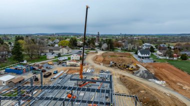 Kırsal alanda bir bina yapım aşamasında. Turnalar ve makineler mevcuttur. Arka planda yakın evler ve açık alanlar görülebilir. Hava bulutlu görünüyor..