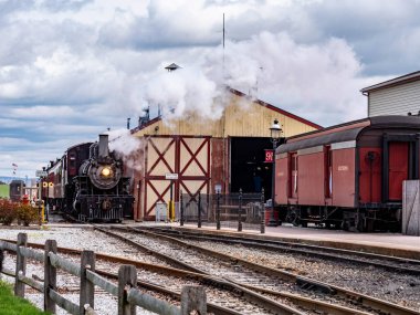 Bir buhar treni eski tren vagonlarının yakınındaki istasyona girer. Hava bulutlu ve motordan buhar yükseliyor. Bölge yolcuların trene binmesi için hazırlandı..