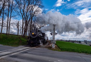 Bir buhar treni tarım arazisi yakınlarındaki tren rayları boyunca hareket eder. Ağaçları geçerken duman çıkarır ve çapraz sinyal verir. Sahne tarlaları ve bulutlu gökyüzünü gösteriyor..
