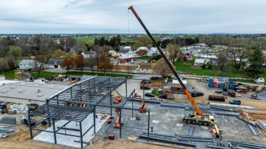 İşçiler, vincin çelik kirişleri kaldırdığı bir inşaat alanında çalışıyorlar. Mekanizma yakınlarda park halinde ve manzara arka planda evleri ve tarlaları gösteriyor..