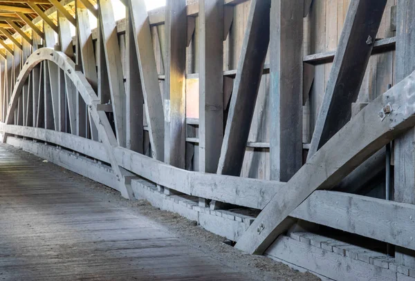 The image shows a wooden bridge with a curved structure inside a covered walkway. Sunlight enters through the openings. The setting appears rural and features natural wood.