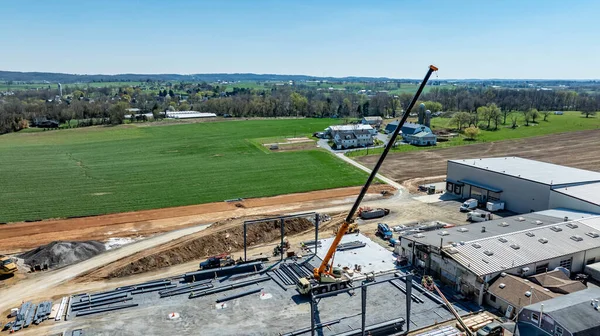İşçiler kırsal bölgedeki bir inşaat sahasına çelik bir yapı inşa ediyorlar. Vinçler ve makineler malzemeleri kaldırmak için kullanılır. Yeşil alanlar ve konut binaları yakınlarda..