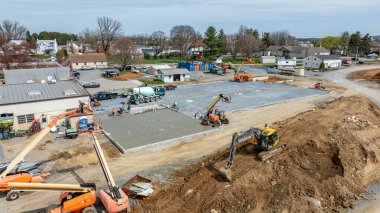 İşçiler betonun döküldüğü bir inşaat sahasında meşguller. Çeşitli makineler çalışıyor. Olay yeri, evlerin ve ekipmanların bulunduğu gelişmekte olan bir alanı gösteriyor..