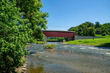 Ağaçlar ve tepelerle çevrili akan bir nehrin üzerinden geçen örtülü bir köprü. Sahne açık mavi gökyüzünü ve yeşil çimenleri gösteriyor. Bu da doğada parlak ve sıcak bir günü gösteriyor..