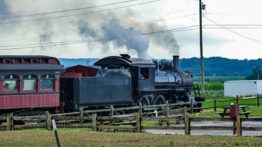 Buhar treni, eski bir yolcu vagonunu yeşil tarlalardan geçirir. Pist boyunca ilerlerken motordan duman yükseliyor. Gökyüzü bulutlu ve manzara kırsal..