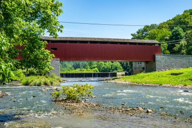 Kırmızı kaplı bir köprü akan bir nehrin üzerinden geçer. Nehir kıyısında yeşil ağaçlar var. Gökyüzü bulutsuz ve berrak. Sahne doğa ve yapıyı kırsal bir ortamda gösteriyor..