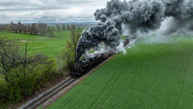 Bir buhar treni yeşil alanlarda yol alır. Kalın siyah duman motordan gökyüzüne yükseliyor. Ağaçlar arka planda ve bulutlar bu bulutlu günde gökyüzünü dolduruyor..