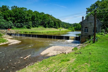 Yeşil kırsal bir alanda taştan bir binanın yakınındaki bir vadi üzerinden bir nehir akar. Elektrik hatları arazide akıyor. Ağaçlar açık mavi gökyüzünün altında uzanır..