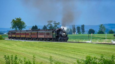 Bir buhar treni, açık yeşil alanlarda seyahat ederken duman üreterek raylar boyunca hareket eder. Ağaçlar arka planda uzak bir kasabayla görülebilir. Gökyüzü açık..