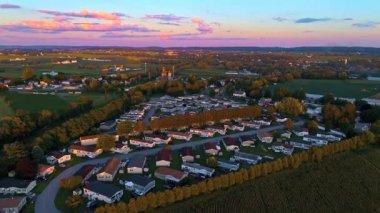 Gün batımı, tarlalarla çevrili küçük bir kasabada üretilen evleri aydınlatıyor. Hava kararırken gökyüzü parlak renkler gösterir. İnsanlar bahçelerde rahatlar ve manzaranın tadını çıkarırlar..