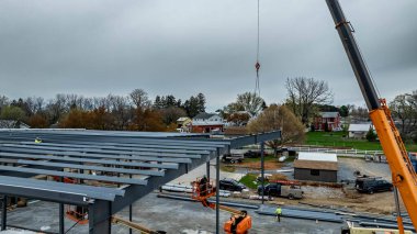 İşçiler kırsal bir yerde yeni bir bina için çelik iskelet monte ediyorlar. Ekipman ve araçlar yakınlarda olduğu için vinç bölümleri kaldırıyor. Gökyüzü bulutlu ve gri.