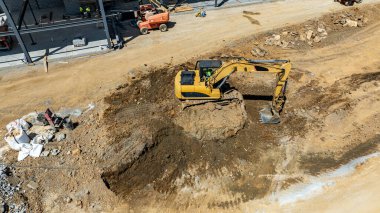 Bir şantiyede inşaat için ağır makineler kullanılıyor. İşçiler burada ve araçlar yakınlarda park halinde. Toprak düz değil, toprak ve kaya yığınları var..