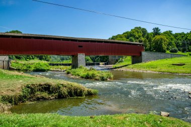 Kapalı bir köprü, yemyeşil tarlalar ve ağaçlarla çevrili akan bir dereyi geçer. Açık mavi gökyüzü ile güneşli bir gün. Su köprünün altından ve küçük bir barajın üzerinden yavaşça akar..