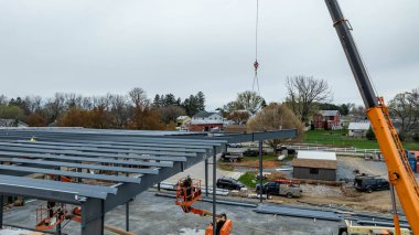 İnşaat işçileri şehre çelik kiriş takmak için makineler kullanıyorlar. İşçiler asansördeyken vinç kirişi kaldırır. Sahne, arka planda aktiviteleri ve binaları gösteriyor..