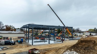 İşçiler bir kasabadaki bir inşaat alanında mevcutlar. Bir vinç yeni bir bina inşa etmek için çelik kirişleri kaldırıyor. Kamyonlar ve makineler yakınlarda park halinde. Gökyüzü bulutlu..