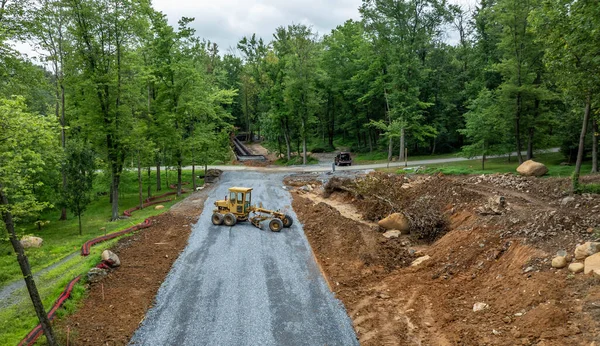 İşçiler ormanlık alanda yol yapmak için makineler kullanıyorlar. Zemin düzleştiriliyor ve ön planda çakıllar var. Bölgenin etrafını saran ağaçlar ve uzaklardan görünen bir patika..